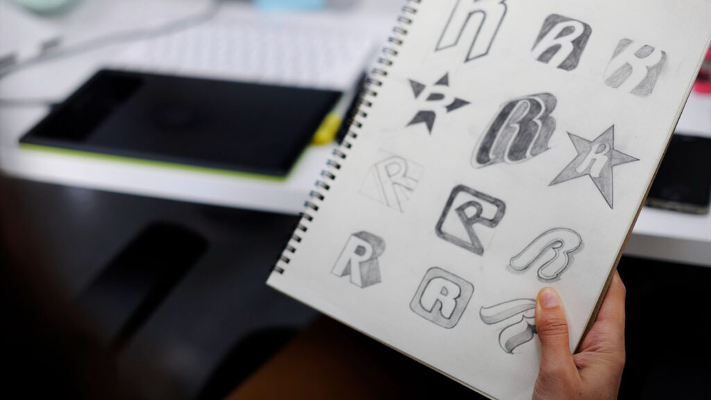 Eine Person hält ein Skizzenbuch mit verschiedenen handgezeichneten Logodesigns, vor allem mit stilisierten „R“-Buchstaben und Sternformen. Im Hintergrund deuten ein Tablet und eine Tastatur auf dem Schreibtisch auf eine nahtlose Website-Navigation hin.
