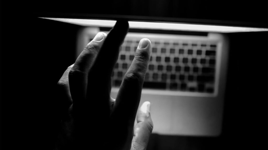 Eine Hand, die in einer dunklen Umgebung nach einem teilweise geschlossenen Laptop greift, wobei die Tastatur durch das Bildschirmlicht schwach beleuchtet wird, suggeriert den Beginn einer nahtlosen Website-Navigation.